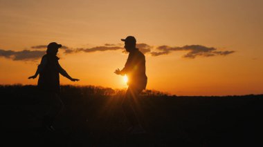 İki çiftçi bir tarlada neşeyle dans ediyor. Siluetler güneşin battığı gökyüzüne karşı.