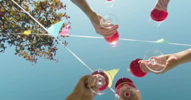 Açık bir gökyüzü altında altı kişi bayrak ve günışığı yapraklarıyla çevrili kırmızı içeceklerle kadeh kaldırıyor. Yüksek kalite fotoğraf