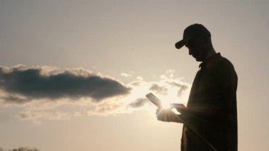 Tarlada, tablet tutan genç bir çiftçinin silueti güneş arka planda batarken çalışıyor.