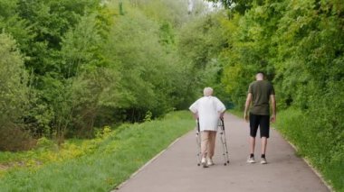 Genç bir adam, yeşil bir parkta, yeşil bir yolda yürüyen yaşlı bir kadını kullanıyor. Destek ve nesiller arası bağlantıyı vurguluyor..
