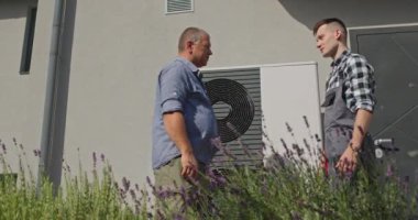 Bir teknisyen ve ev sahibi, bir banliyö ortamında harici bir ısı pompası ünitesinin yanında el sıkışarak bakım ya da kurulum hizmetinin tamamlanmasına işaret ederler.. 