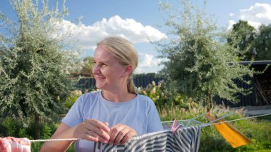 Güneşli bir bahçede çamaşır ipine çamaşır asarken gülümseyen orta yaşlı bir kadın, güzel bir günde ev işlerinin keyfini çıkarıyor. Yüksek kalite fotoğraf