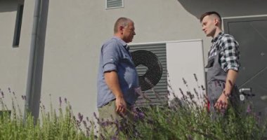 Bir teknisyen ve ev sahibi, bir banliyö ortamında harici bir ısı pompası ünitesinin yanında el sıkışarak bakım ya da kurulum hizmetinin tamamlanmasına işaret ederler.. 