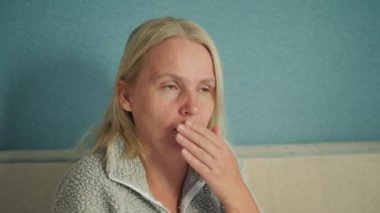 Orta yaşlı bir kadın yatakta oturuyor, boğaz ağrısı çekiyor, lenf bezlerine acı dolu bir ifadeyle masaj yapıyor.