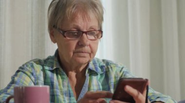 Üst düzey bir kadın mutfak masasında akıllı telefon kullanıyor, bir fincan sıcak çayın yanında oturuyor, rahat ve sade bir atmosfer yaratıyor. Yüksek kalite 4k görüntü