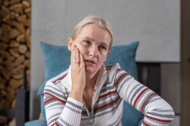 Middle-aged woman indoors with a pensive expression, resting her hand on her cheek in a cozy home setting.  clipart