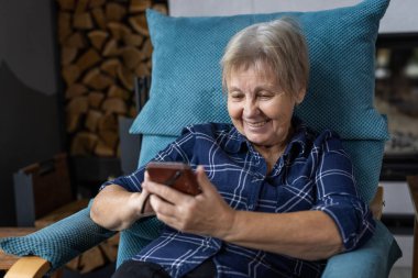 Evde rahat bir koltukta dinlenirken akıllı telefon kullanan gülümseyen yaşlı bir kadın. Yüksek kalite fotoğraf