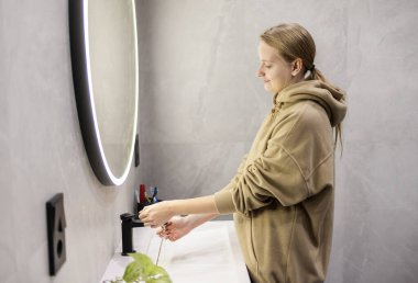 Genç bir kadın ellerini modern bir banyo lavabosunda parlak bir aynayla yıkıyor ve temiz ve minimalist bir atmosfer yaratıyor. Yüksek kalite fotoğraf
