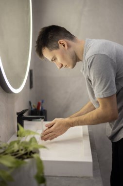 Modern bir lavaboda ellerini yıkayan genç bir adam, arka planda parlak bir ayna ile temizlik ve hijyene odaklanıyor. Yüksek kalite fotoğraf