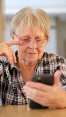 Gözlüklü yaşlı kadın akıllı telefonuna şaşkın şaşkın bakıyor, şaşkın bir ifadeyle konsantre oluyor. Yüksek kalite fotoğraf