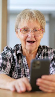 Senior woman looking surprised and delighted at her smartphone, mouth open in awe, wearing glasses and a plaid shirt. High quality photo clipart