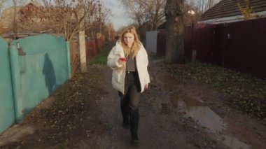 Genç bir kadın, etrafındaki eski ve çürüyen evlerle çamurlu bir yolda yürüyor. Riskli bir mahallede akıllı telefon kullanıyor. 