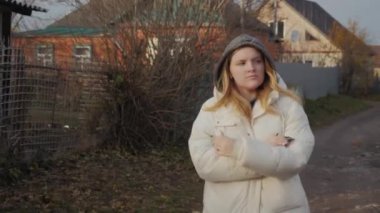 Çamurlu bir sokakta yürüyen genç bir kadın harap olmuş tek katlı evlerle dolu, soğuktan titriyor ve ellerini ısıtıyor.