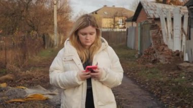 Genç bir kadın, tehlikeli bir mahallede, eski ve harap evlerle çevrili çamurlu bir yolda akıllı telefon kullanarak yürüyor. Yüksek kalite 4k görüntü