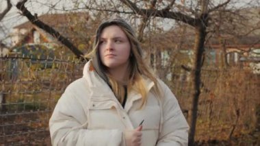 Genç bir kadın soğuk sonbahar rüzgarından titreyen ve ellerini ısıtmaya çalışan eski kırsal evlerin arka planında duruyor. Yüksek kalite 4k görüntü