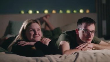 Genç bir çift loş ışıkta bir yatakta oturup arka planda ışıldayan bir çelenkle neşeli filmler izleyip Noel havası yaratıyor. Yüksek kalite 4k görüntü