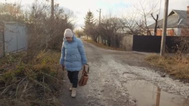 Çamurlu kırsal bir yolda yürüyen yaşlı bir kadın, elinde bir çanta ve bir telefon, sıcak mavi bir ceket ve şapka giymiş, sessiz bir köy kurma ile çevrili