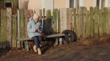 Ukrayna 'nın kırsal bir köyünde, ahşap bir çitin yanındaki bankta cep telefonu kullanan yaşlı bir kadın. Yüksek kalite 4k görüntü