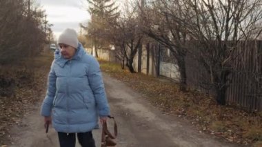 Sessiz bir köyde kırsal bir yolda yürüyen yaşlı bir kadın elinde bir telefon ve bir çanta, sıcacık bir şekilde mavi bir ceket ve şapka giymiş. Yüksek kalite 4k görüntü