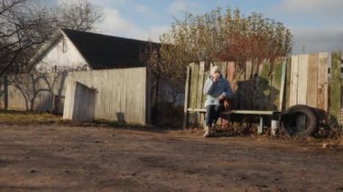 Huzurlu bir kırsal alanda, arkasında yıpranmış ahşap bir çitle kırsal bir bankta otururken yaşlı bir kadın bardaktan su içiyor. Yüksek kalite 4k görüntü