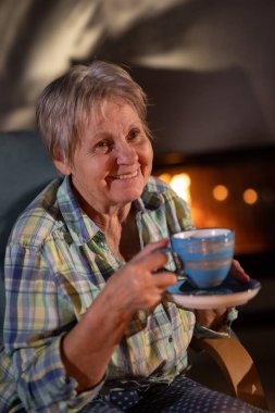 Yaşlı bir kadın şöminenin yanında oturuyor, elinde mavi bir çay fincanı, sıcacık ve rahat bir akşamın tadını çıkarırken sıcacık gülümsüyor. Yüksek kalite fotoğraf