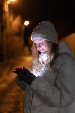 Gece loş bir sokakta yürüyen genç bir kadın, akıllı telefonuna odaklanmış, sıcak giysileri ve sakin, yansıtıcı bir atmosferi var. Yüksek kalite fotoğraf