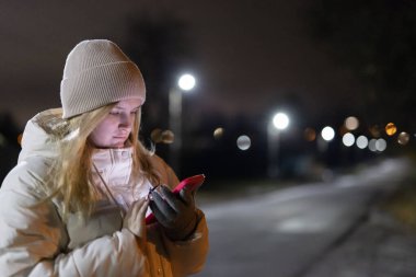 Young woman in a warm jacket and hat using her smartphone outdoors at night, illuminated by streetlights with a blurred background. High quality photo clipart