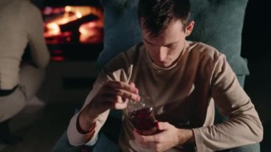 Genç bir adam şöminenin yanındaki koltukta oturmuş elinde bir şişe alkolle arka planda alevlere odun atarken. Yüksek kalite 4k görüntü