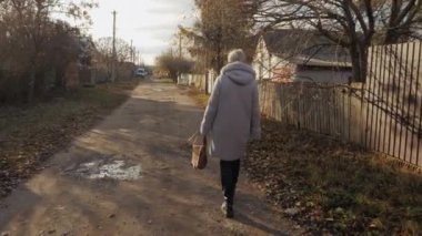  Sıcak sonbahar ceketli bir kadın çamurlu kırsal bir yolda yürüyor, bir çanta taşıyor, yanında mütevazı evler ve yerde don oluşuyor.