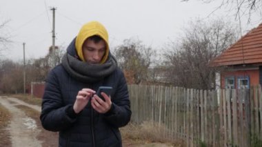 Sıcak ceketli, parlak sarı kapüşonlu genç bir adam Ukrayna 'nın otantik bir köyünde elinde akıllı bir telefonla kırsal bir sokakta yürüyor. Yüksek kalite 4k görüntü