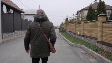 Omuz çantalı genç bir adam sokakta özel evlerin arasında yürüyor, arkadan bakılıyor, bir posta teslimatı konseptini çağrıştırıyor. Yüksek kalite 4k görüntü
