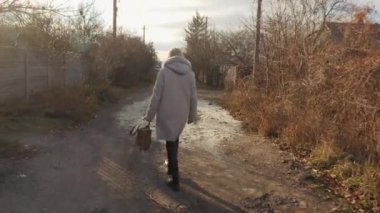 Sonbahar ceketli bir kadın çantasıyla kırsal bir toprak yolda yürüyor, mütevazı evlerle çevrili, su birikintileri ve donmuş zeminle birlikte. Yüksek kalite 4k görüntü