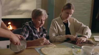 Son sınıf öğrencisi bir kadın, torunlarıyla birlikte parlayan bir şöminenin yanında hamur tabanlı bir yemek hazırlıyor. Birbirine sıkı sıkıya bağlı aile, kış tatili beklentileriyle dolu sıcak ve şenlikli bir atmosferin tadını çıkarıyor.