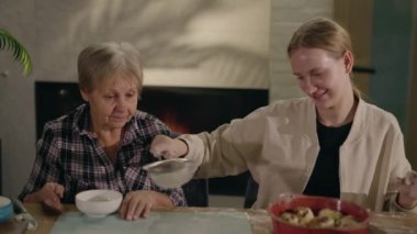 Genç bir kız, yanan bir şöminenin yanındaki masada otururken büyükannesinin pizza için hamur hazırlamasına yardım eder. Birbirine kenetlenmiş aile, yaklaşan kışı bekleyerek sıcacık bir anın tadını çıkarıyor.