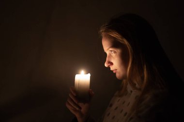 Genç bir kadın, yanan bir muma dikkatle bakar, ılık parıltısı, yüzünü karanlık bir arka plana doğru hafifçe aydınlatır. Sahne, derin düşüncelere dalma ve samimiyet hissi uyandırıyor. Yüksek kalite fotoğraf