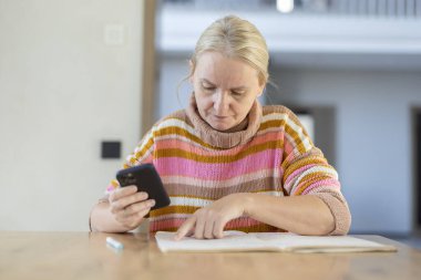 Renkli çizgili süveter giyen orta yaşlı bir kadın elinde akıllı bir telefonla, iyi aydınlatılmış bir odada, ahşap bir masada otururken kitap okuyor. Yüksek kalite fotoğraf