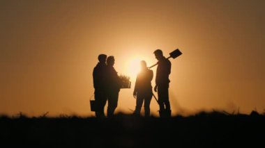 Bir grup çiftçi ellerinde aletler ve bir kutu fidanla gün batımında bir tarlada durup sohbet ediyorlar..