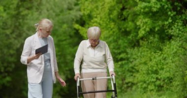 Kendini adamış bir bakıcı yeşil park ortamında yürüyen bir kadına destek oluyor.