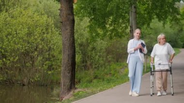 Orta yaşlı bir kadın, yemyeşil bir parkta yürürken yürüteç kullanan yaşlı bir kadına yardım ediyor. Sahne, bakım, arkadaşlık ve açık hava etkinliğinin önemini vurguluyor.