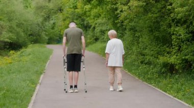 Yürüteç kullanan genç bir adam ve yaşlı bir kadın yemyeşil bir parkta yol boyunca yan yana yürüyorlar. Sahne desteğin, nesiller arası bağın ve faydaların önemini vurguluyor.