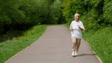 Yaşlı bir kadın güzel, yemyeşil bir park içinde manzaralı bir yolda ferahlatıcı bir koşunun tadını çıkarıyor.