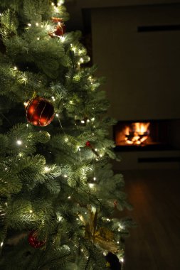 Güzel dekore edilmiş bir Noel ağacı, kırmızı süslemeleri ve parıldayan ışıklarıyla, rahat bir oturma odasında, arka planda parlayan bir şömineyle.