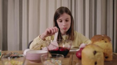 Genç bir kız Paskalya yumurtalarını canlı yumurtalar ve taze pişmiş Paskalya kekleri de dahil olmak üzere bayram eşyalarıyla dolu bir masada boyamaya konsantre oluyor. Yüksek kalite 4k görüntü