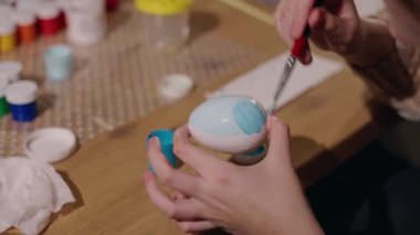 Elleri yakın plan, açık mavi ve beyaz renkli bir Paskalya yumurtası boyuyor. Etrafı fırçalarla çevrili ve ahşap bir masaya kavanozlar boyuyor. Yüksek kalite 4k görüntü