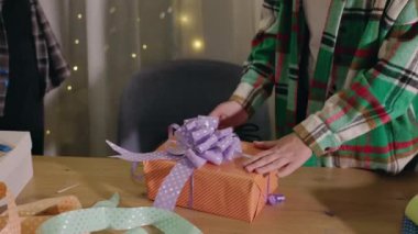 Turuncu bir hediye kutusuna mor benekli bir kurdele bağlayan ellerin yakın çekimi. Etrafı renkli kurdelelerle çevrili. Ahşap bir masa ve şenlik ortamı. Yüksek kalite 4k görüntü