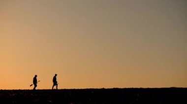 Işıl ışıl güneşe karşı siluet oluşturan iki çiftçi, gün doğumunda ya da batımında açık bir arazide yürüyorlar, tarım aletleri taşıyorlar. Huzurlu sahne, kırsal yaşamın özünü ve adanmışlığı yansıtıyor.