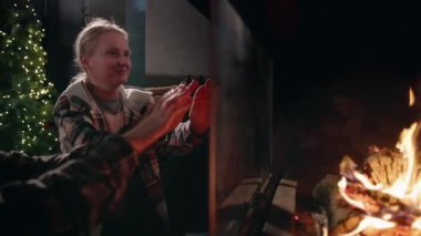 Bir kadın ellerini modern bir şöminenin yanında ısıtıyor, arka planda süslü bir Noel ağacı olan festival odasının rahat atmosferinde gülümsüyor. Yüksek kalite 4k görüntü