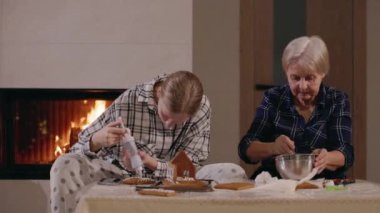 Farklı nesillerden üç kadın sıcak bir şöminenin yanında zencefilli kurabiye evi dekore ederek neşeli bir bayram geleneğini paylaşıyorlar. Yüksek kalite 4k görüntü