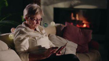 Rahat bir kanepede, şömine ve dekoratif ışıklarla aydınlatılmış rahat bir oturma odasında akıllı telefon kullanan son sınıf kadını. Yüksek kalite 4k görüntü
