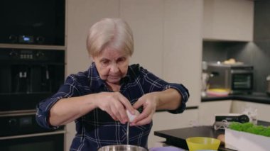 Modern bir mutfakta yaşlı bir kadın yemek pişirme işine odaklanmak için yumurtayı dikkatlice bir kaseye kırıyor. Yüksek kalite 4k görüntü